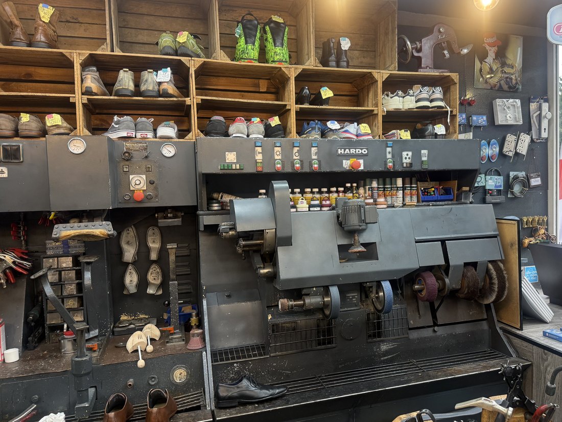 Die Ausputzmaschine in der Werkstatt, darüber Holzkisten mit fertigen und wartenden Schuhen.